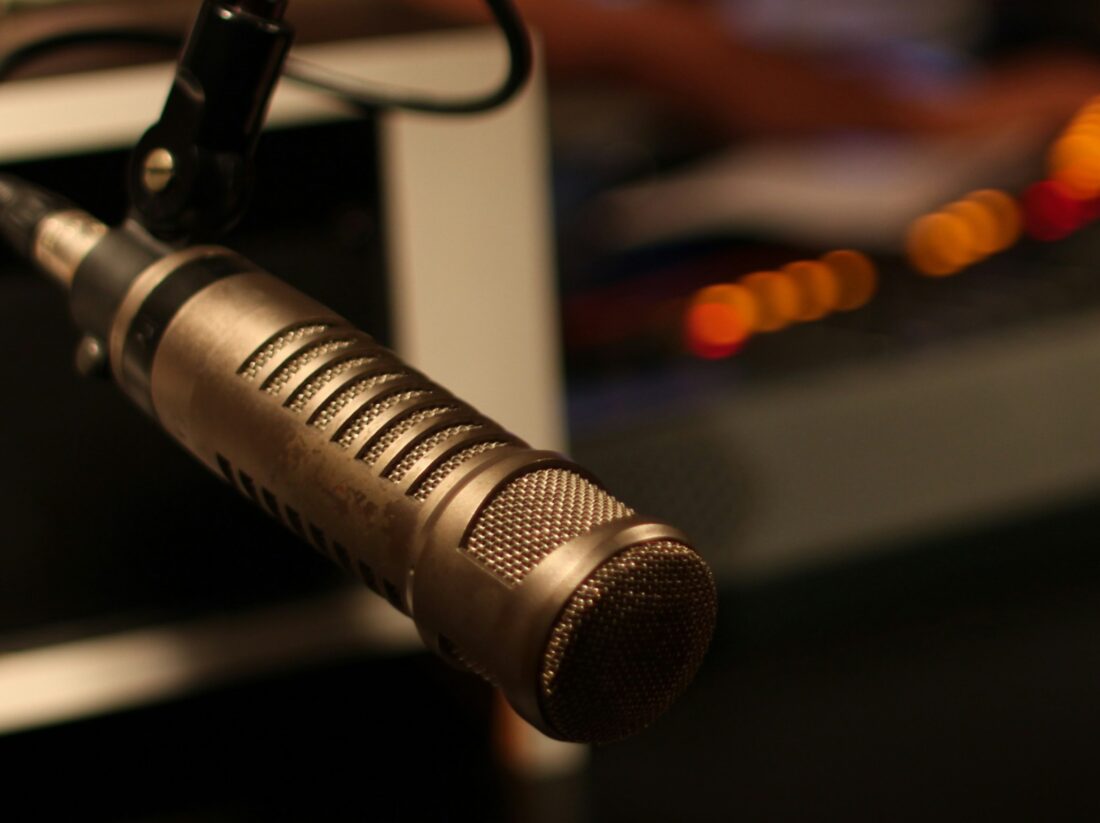 Microphone professionnel dans un studio de radio, entouré d’un éclairage tamisé et d’un arrière-plan flou où l’on devine le matériel de diffusion.